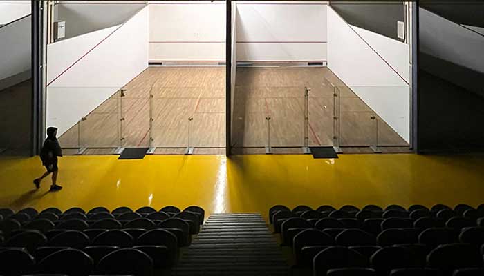 An empty squash court awaits play as a person walks past the seating area inside an indoor sports arena. — Unsplash