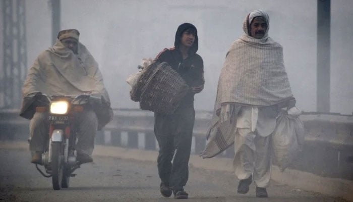 A motorcyclist passes by men walking along a road in dense fog on a cold day in Lahore. — Reuters/File