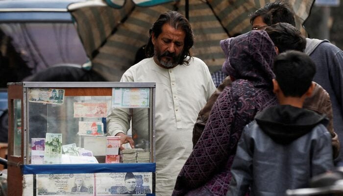 A currency broker stands near his booth, which is decorated with pictures of currency notes, while dealing with customers, along a road in Karachi. — Reuters/File