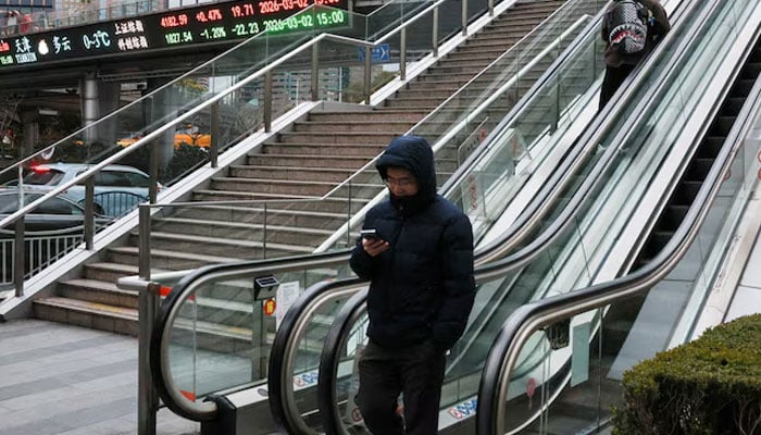 An electronic board shows Shanghai stock indices as people ride an escalator on a pedestrian bridge in the Lujiazui financial district in Shanghai, China, March 2, 2026. — Reuters