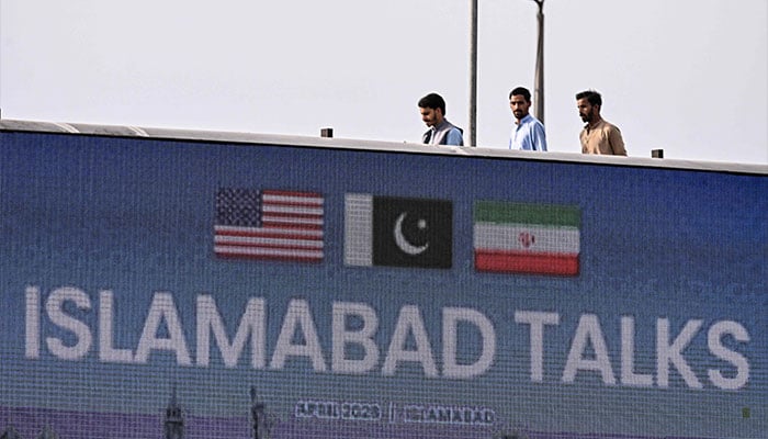 Youth walk behind a digital screen displaying news of US-Iran peace talks along a road in Islamabad on April 10, 2026. — AFP