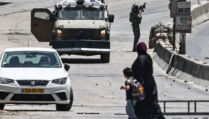 A woman and a child cross a street as Israeli security forces patrol during a military raid in the Qalandia refugee camp, south of Ramallah in the Israeli-occupied West Bank, on May 11, 2026. —AFP