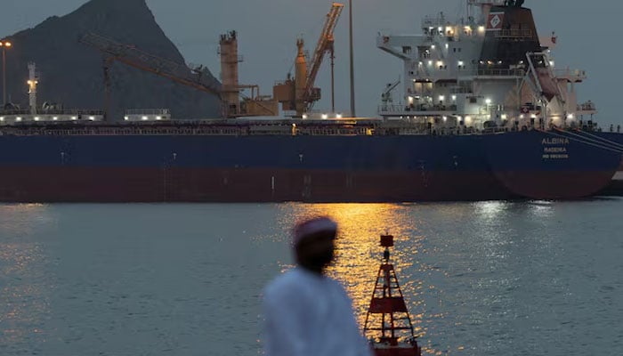 A man sits on the waterfront as a vessel sits at anchor inside Sultan Qaboos Port, amid the U.S.-Israeli conflict with Iran, in Muscat, Oman, March 20, 2026. — Reuters