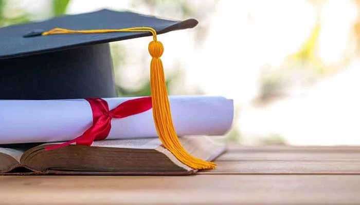 The representational image displays a graduation cap (also known as a mortarboard) with a yellow tassel, placed on top of an open book and a rolled-up diploma tied with a red ribbon. — Unsplash/File