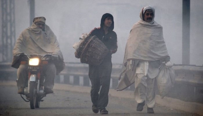 A motorcyclist passes by men walking along a road in dense fog on a cold day in Lahore. — Reuters/File