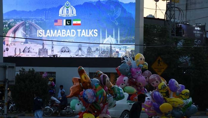 A balloon seller walks near a screen with an image referring to the US-Iran peace talks on April 11, 2026. — Reuters
