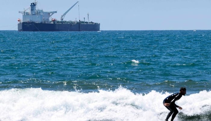 A surfer rides a wave in front of the Sea Voyager crude oil tanker, anchored in the Pacific Ocean off the coast of the Chevron El Segundo refinery, on April 23, 2026, in El Segundo, California. — AFP