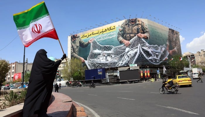 Above, a woman holds Irans national flag while standing near a billboard with a sentence reading The Strait of Hormuz remains closed at the Enqelab Square in Tehran, on April 5, 2026. — AFP